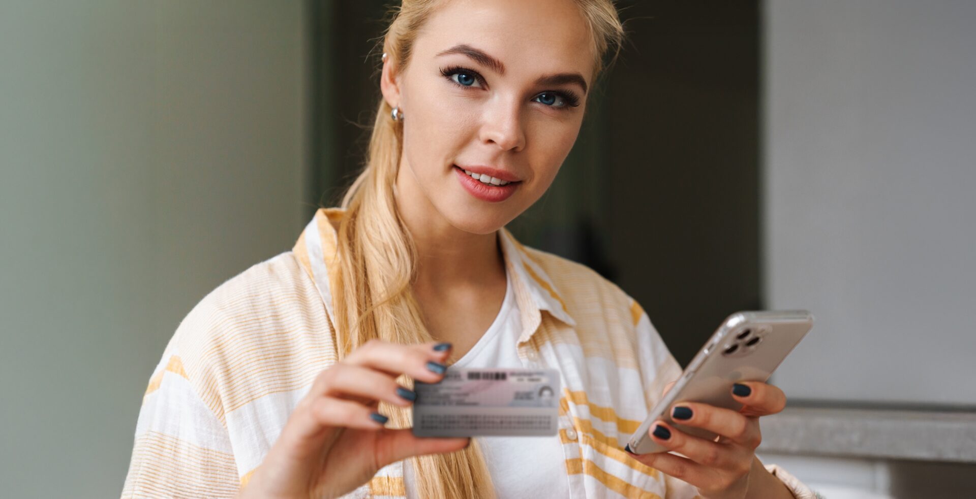 Kobieta z długimi blond włosami patrzy w obiektyw i uśmiecha się, trzymając w jednej ręce smartfon, a w drugiej plastikowy dowód osobisty. Wygląda, jakby korzystała z aplikacji mobilnej lub logowała się online przy użyciu dokumentu tożsamości.