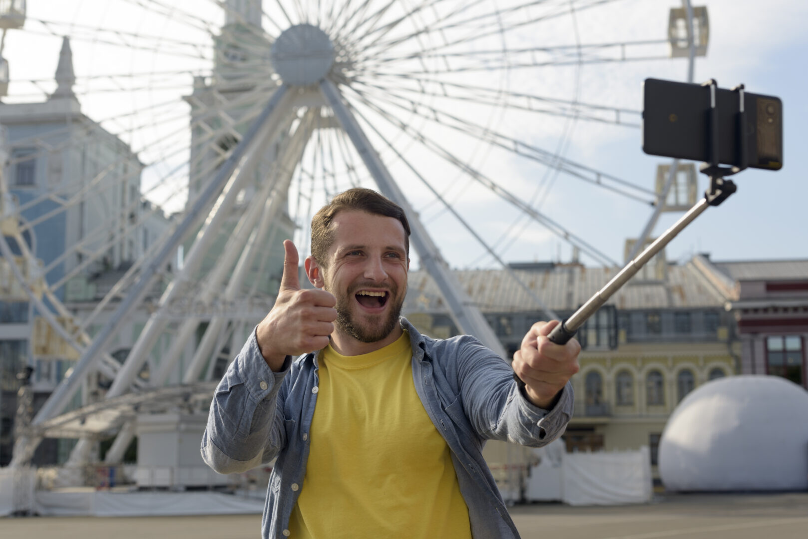 Mężczyzna stoi przed dużym diabelskim młynem, trzymając kijek do selfie z telefonem i pokazując kciuk w górę. Uśmiecha się szeroko, sprawiając wrażenie zadowolonego i pełnego energii, prawdopodobnie robiąc zdjęcie lub nagranie z wyjazdu.