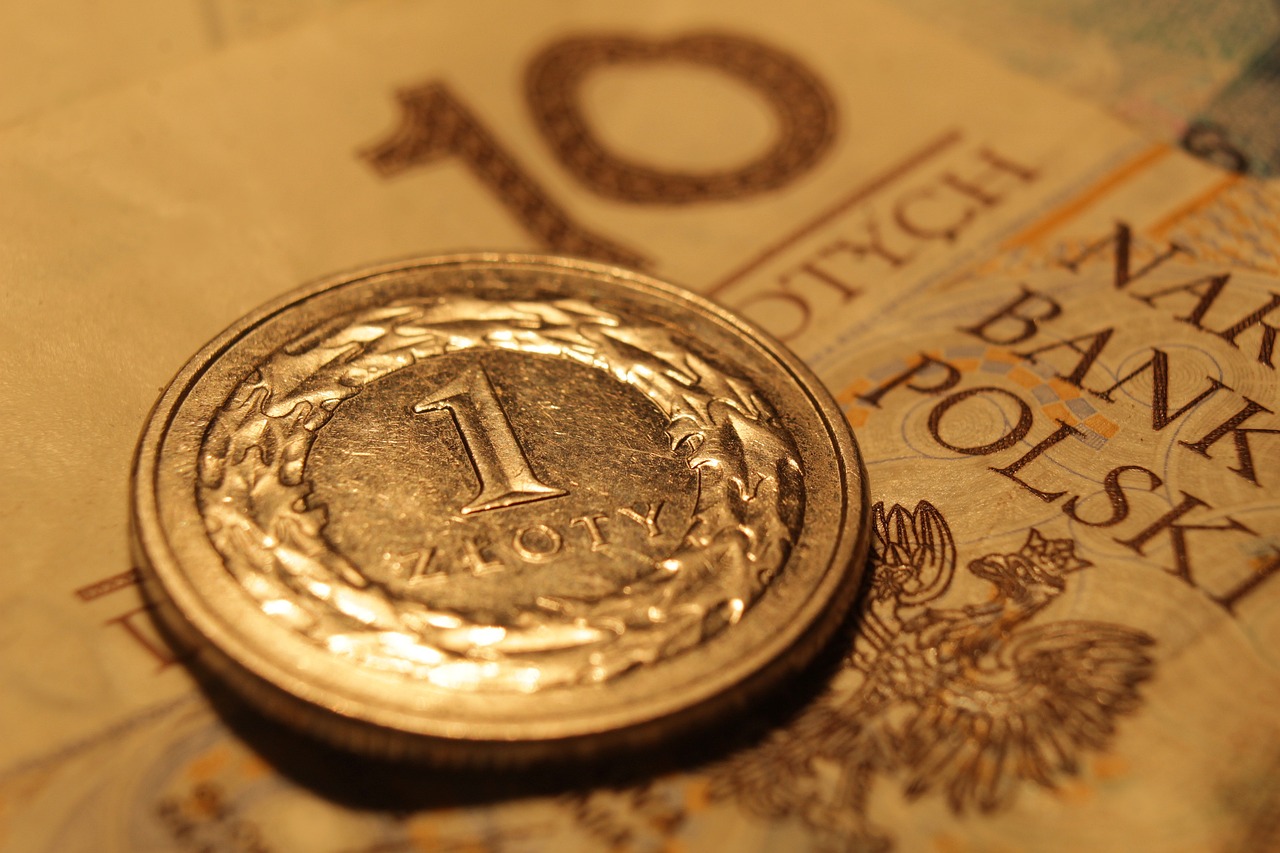 Zbliżenie monety o nominale 1 złoty leżącej na banknocie 10 złotych. Na banknocie widoczne są fragmenty napisu „Narodowy Bank Polski” oraz godło Polski z orłem.