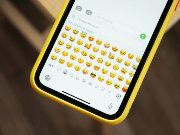 Zbliżenie smartfona w żółtym etui z otwartą klawiaturą emoji. Na ekranie widać różne emotikony – uśmiechy, mrugnięcia, serduszka i inne wyrażające emocje