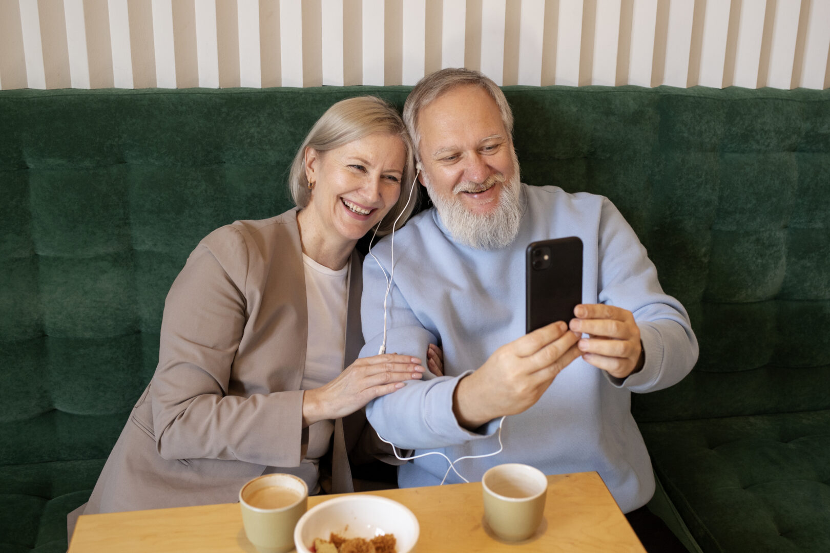 grupa rodzinna w komunikatorze. Starsze małżeństwo siedzi razem na zielonej kanapie, uśmiechając się do telefonu, którym robią selfie lub prowadzą rozmowę wideo. Mają wspólne słuchawki, a na stole przed nimi stoją filiżanki z kawą i miseczka z ciastem. Scena emanuje ciepłem i radością.