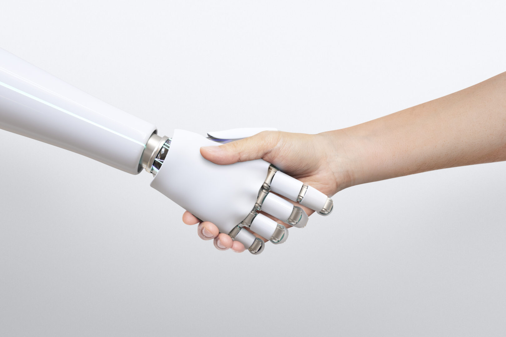 Na zdjęciu widać uścisk dłoni człowieka i robota. Ludzka ręka ściska mechaniczną, białą dłoń robota na neutralnym tle. Obraz symbolizuje współpracę człowieka z technologią, zaufanie do sztucznej inteligencji i harmonię między ludzkim światem a światem maszyn.