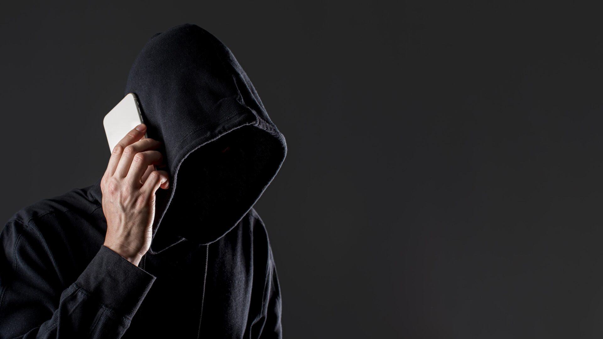 Na zdjęciu widać osobę w czarnej bluzie z kapturem, trzymającą telefon przy uchu. Twarz jest ukryta w cieniu, co tworzy atmosferę tajemniczości i zagrożenia. Obraz może symbolizować oszusta telefonicznego (vishing) lub cyberprzestępcę próbującego wyłudzić dane.