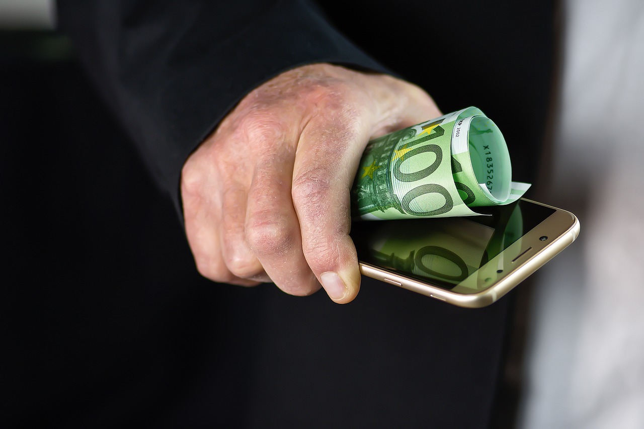 Zbliżenie dłoni trzymającej smartfon oraz zwinięty banknot o nominale 100 euro. Obraz symbolicznie przedstawia powiązanie technologii mobilnych z finansami lub płatnościami elektronicznymi.